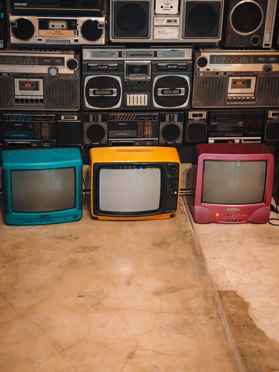 Three brightly colored portable televisions in teal, yellow and red, with retro radios stacked behind them
